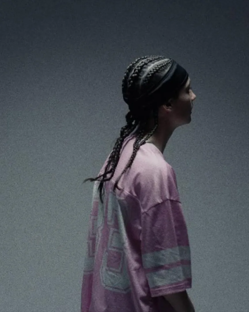 Oasky de perfil, con trenzas y camiseta rosada, sobre fondo gris